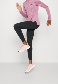 Camicia rosa chiaro a maniche lunghe con colletto a mezza zip, leggings neri e scarpe da ginnastica rosa chiaro con dettagli bianchi e gialli.