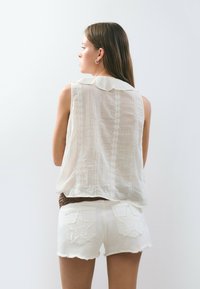 Blusa blanca sin mangas ligera con un cuello con volantes y bordados intrincados, combinada con pantalones cortos blancos desgastados que presentan bolsillos en forma de estrella.