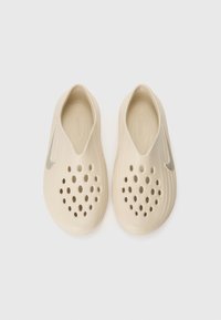Des chaussures beiges à enfiler en matériau doux avec des trous de ventilation, arborant un accent argenté du logo Nike sur le côté.
