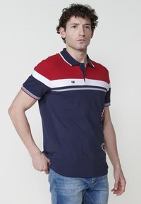 Mornarska polo majica s crveno-bijelim prugama, kratkim rukavima, ovratnikom i logotipom na prsima; sadrži stilizirani rub i dekorativni bočni detalj.