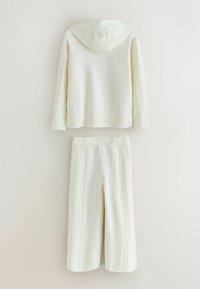 Balta adīta kapuce ar kapuci un ribotu apakšmalu. Apvienota ar atbilstošām biksēm, kurām ir teksturēts kabelis gar malām.