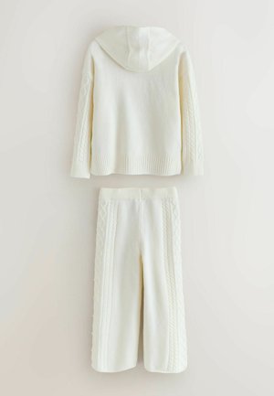 Balta megzta kapučinė striukė su kapišonu ir rilstukais apačioje. Derinama su atitinkamomis kelnėmis, turinčiomis tekstūruotą kabelių raštą šoninėse dalyse.