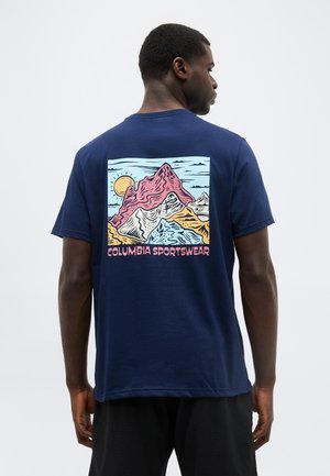Άνδρας που φορά μπλε ναυτικό T-shirt με πολύχρωμο γραφικό τοπίου ορεινών και ήλιο, και κείμενο "Columbia Sportswear" στο πίσω μέρος.