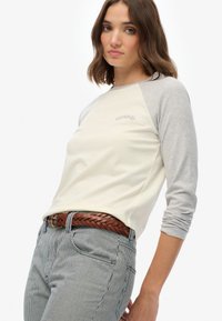 Młoda kobieta z brązowymi włosami do ramion, ubrana w kremową i szaro-długą bluzkę oraz pasiaste spodnie z plecionym brązowym paskiem.