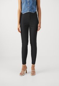 Zwarte slim-fit broek met een gladde textuur, hoge taille en zonder zichtbare sluitingen, gecombineerd met sandalen met hakken en bandjes.
