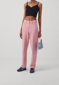 Jean taille haute rose clair, coupe décontractée, associé à un crop top noir à bords festonnés. Un petit sac à main bleu clair est tenu. Des baskets décontractées complètent le look.
