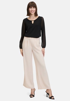 Schwarze, halbtransparente Bluse mit langen Ärmeln und Schlüssel-ausschnitt, kombiniert mit hellbeigen, weitem Hosen. Schwarze, hohe Sandalen vervollständigen das Outfit.