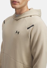 Sudadera beige hecha de tela suave, con un bolsillo frontal, capucha con cordón y líneas de acento negras en los hombros. Logotipo de Under Armour visible.