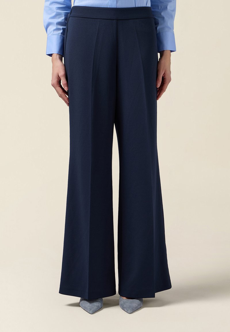 Persona con pantalones anchos color azul marino con pliegues frontales y zapatos azules de punta puntiaguda, de pie frente a un fondo neutro.