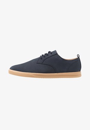 Zapato informal azul marino con cordones, suela de goma color tostado y lengüeta para tirar en el talón, mostrado de lado sobre un fondo blanco.