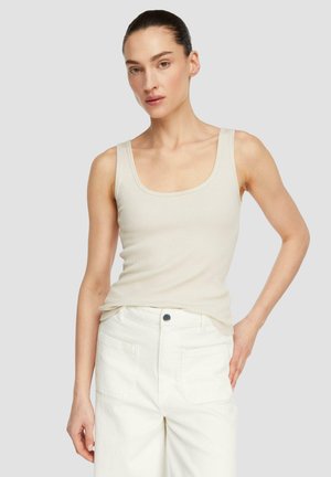 Frau mit dunklem Haar in einem niedrigen Dutt, trägt ein beiges, geripptes Tanktop und weiße, hochgeschnittene Hosen, steht mit einer Hand auf der Hüfte.