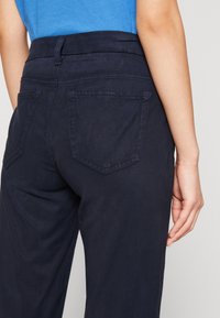 Navy blue housut, joissa on istuva vyötärö, kaksi takataskua ja hienovarainen ommelarkkitehtuuri. Kangas näyttää sileältä ja siinä on kevyt kiilto.