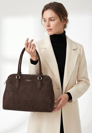 Vrouw met een beige jas en een zwarte coltrui houdt een bruine leren handtas met metalen rits en dubbele hengsels vast.