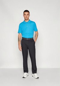 Polo azul com três botões, combinado com calças pretas feitas à medida. Modelo usa ténis brancos; design minimalista e tecido leve.