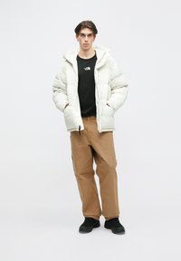 Doudoune blanche avec capuche, fermeture éclair et poches latérales, portée sur un t-shirt noir, accompagnée de pantalons beiges et de chaussures noires.