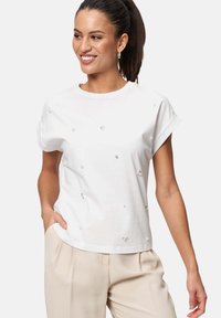 T-shirt blanc à manches courtes en coton avec des embellissements argentés dispersés. L'encolure est de style ras du cou et la coupe est décontractée.