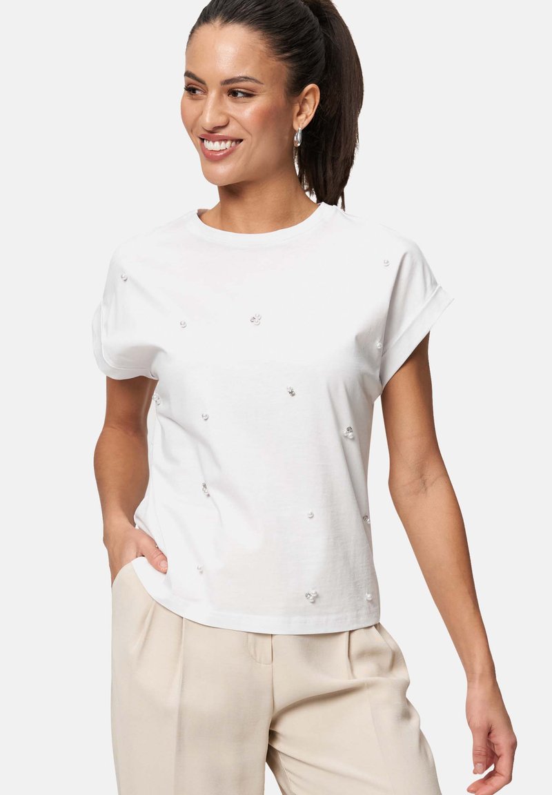 T-shirt blanc à manches courtes en coton avec des embellissements argentés dispersés. L'encolure est de style ras du cou et la coupe est décontractée.