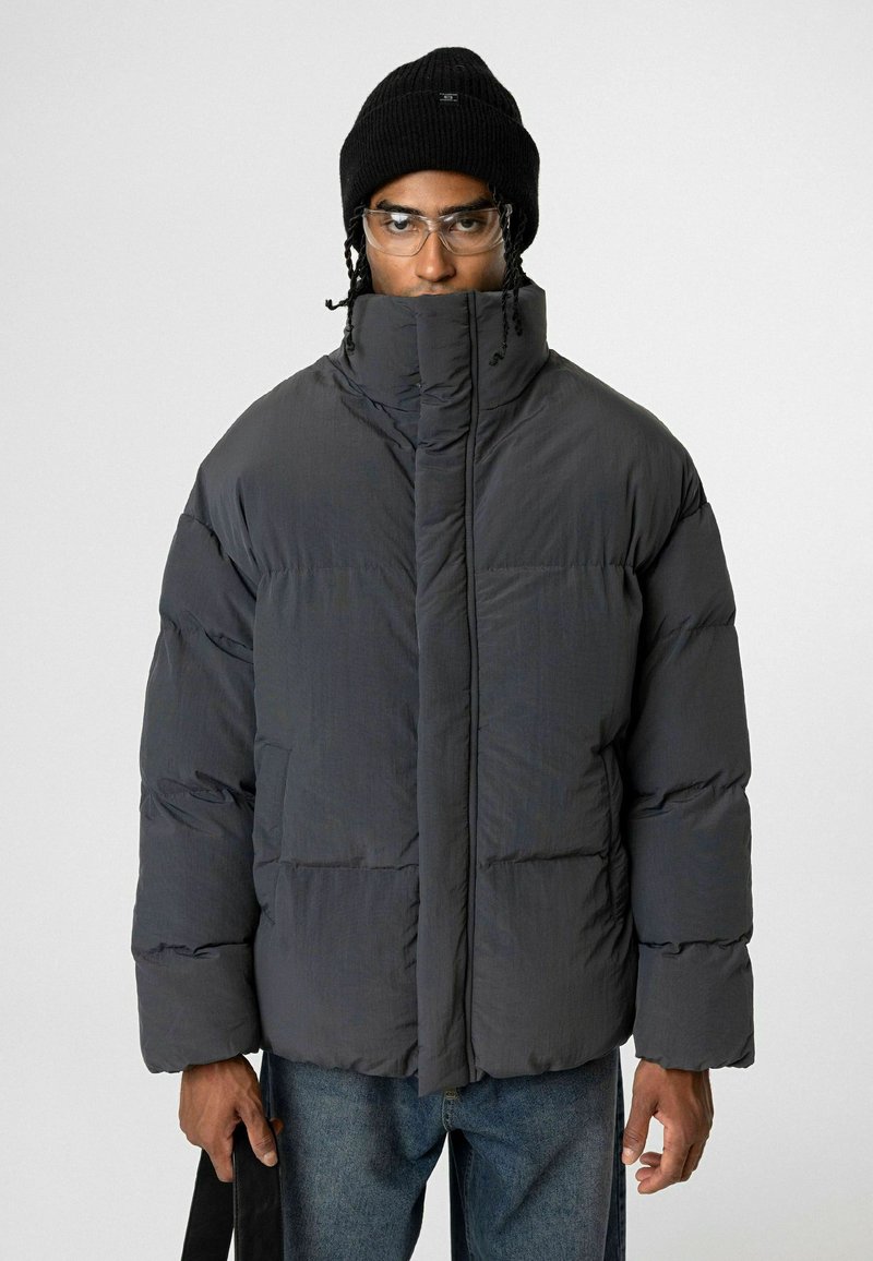 Donkergrijze gewatteerde jas met hoge kraag, gestikte design en oversized pasvorm. Gecombineerd met een zwarte beanie en een bril, vastgehouden door een model.