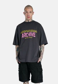 Camiseta extragrande de color gris oscuro con la palabra "ARCHIVE" en rosa, acompañada de texto adicional en amarillo. Llevada con pantalones cortos de cargo negros, mostrada en una persona tatuada.