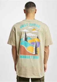 Beige suuri t-paita, jossa on monivärinen seikkailuteemainen graafinen kuvaus, jossa on puita, vuoria, pilviä ja aurinko selässä.
