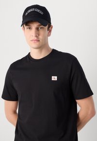Zwarte katoenen T-shirt met ronde hals, met een klein rood en wit Calvin Klein-logo op de linkerkant van de borst. Draagt een zwarte pet.
