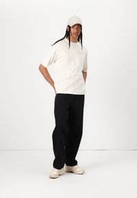 Stribet oversized t-shirt i crema med tynde orange linjer, parret med sorte brede bukser og beige sneakers. Kappen er lys grå.