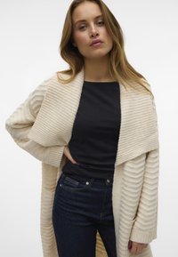 Kremasti rebrasti oversized kardigan s šalom, nosi se preko crnog topa, uparen s tamnim trapericama. Vidljivi su teksturirani pleteni detalji.