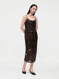 Femme portant une robe midi noire en voile avec des paillettes et de fines bretelles, associée à des chaussures noires à talons pointus, debout les mains derrière le dos.