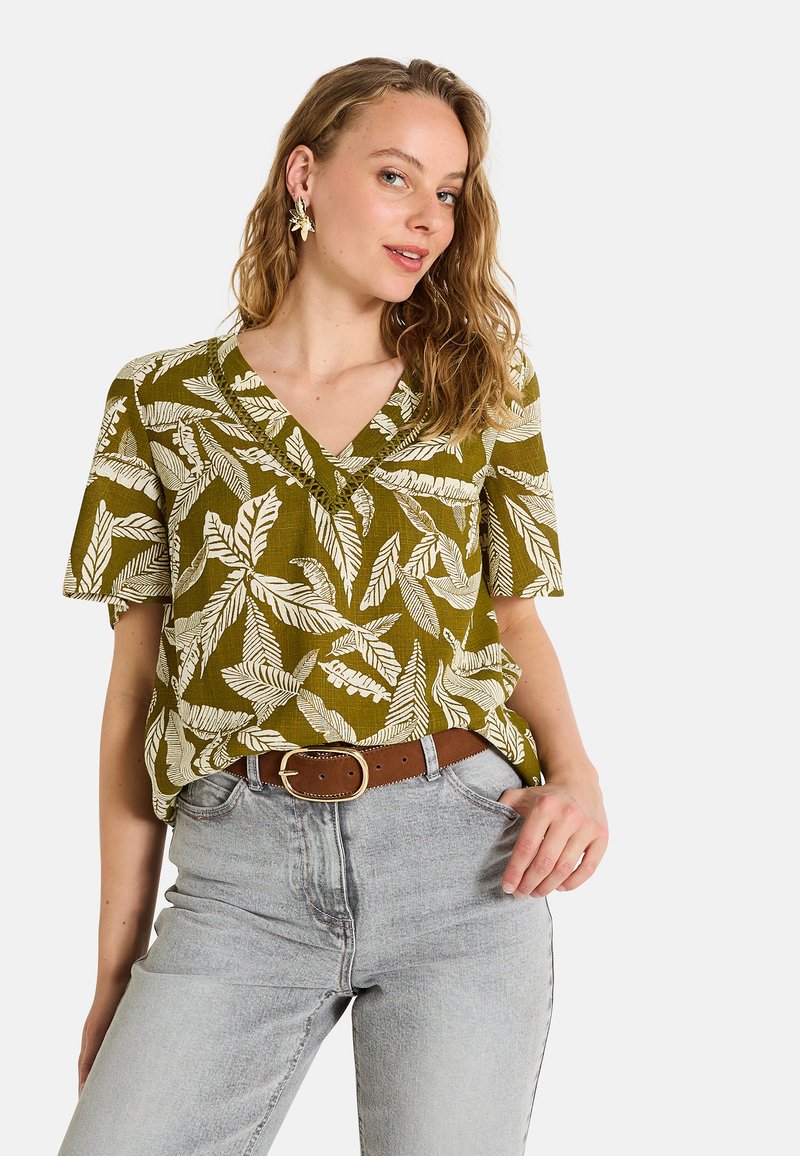 Chemise à manches courtes vert olive avec des motifs de feuilles blanches, décolleté en V et tissu texturé, associée à un jean en denim clair taille haute et une ceinture marron.