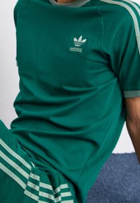 Grön bomulls T-shirt med rund halsringning och grå detaljer, med en broderad Adidas-logotyp på bröstet, matchad med randiga byxor.