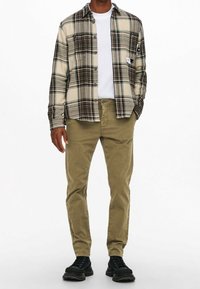 Flannel skjorte i beige med brun og grøn ternet mønster, knappet, over en hvid t-shirt, parret med olivengrønne bukser og sorte sko.