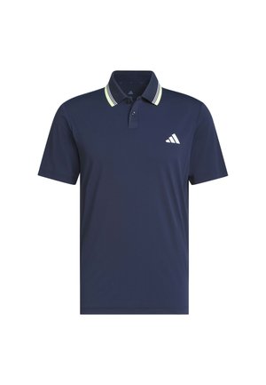 Homme portant un polo bleu marine avec un col à rayures blanches et un pantalon jaune clair, debout avec une main dans la poche sur un fond uni.