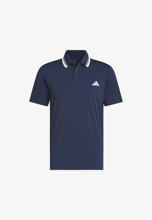 Mann trägt ein marineblaues Poloshirt mit weiß gestreiftem Kragen und hellgelber Hose, steht mit einer Hand in der Tasche vor einfarbigem Hintergrund.