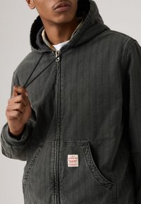 Dark gray hooded jacket with vertical stripes, a front pocket, and a label featuring red text. Material appears textured and durable.