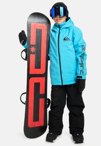 Veste imperméable bleue avec des accents noirs et un logo, associée à un pantalon isolant noir. Tenant un snowboard noir avec des graphismes rouges.