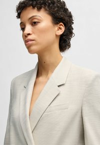  licht beige blazer van een gestructureerde stof, met notch-lapel, enkele borstzak aan de linkerkant en een getailleerde fit.