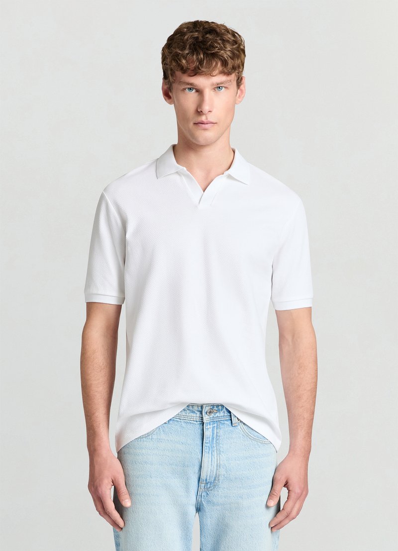 Polo blanc en tissu texturé, avec un col et des manches courtes, associé à un jean bleu clair. Design simple et couleur neutre.