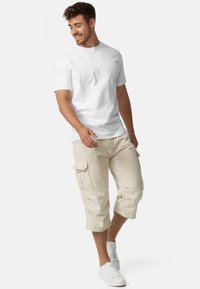 T-shirt en coton blanc associé à un short cargo beige avec des poches latérales. Le modèle porte des baskets blanches, illustrant une tenue décontractée d'été.