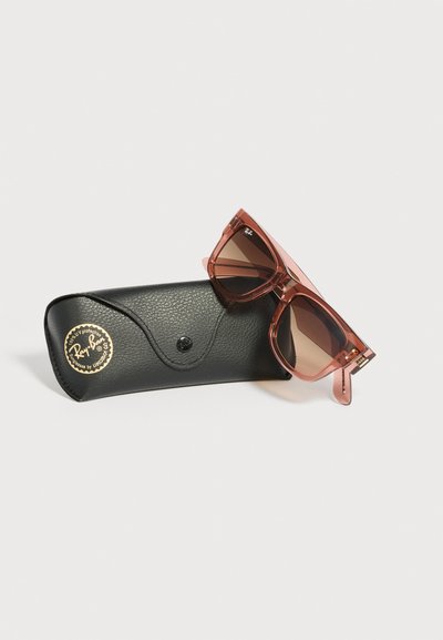 Розови полупрозрачни слънчеви очила Ray-Ban с квадратна рамка, поставени върху черен кожен калъф с черно и златно лого на Ray-Ban.