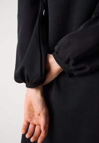 Zwarte jurk met lange, opgezette mouwen en elastische manchetten. Close-up van de achterkant van de hand tegen het lichaam. Glad stoffen textuur.