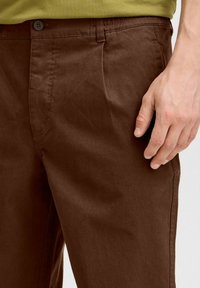 Braune Baumwollhosen mit glatter Textur, ausgestattet mit Vorderaschen, einem Knopfverschluss und sichtbaren Nähdetails. Die Hand ist in der Nähe der Taille platziert.