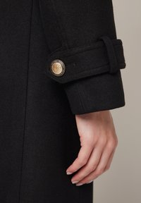 Czarny wełniany rękaw płaszcza z detalem guzika z złotym emblematem, gładka tekstura oraz kontrastujący pasek na mankiecie.