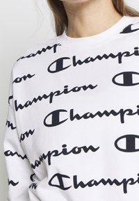 Biała bluza z powtarzającym się czarnym napisem "Champion" i logo, noszona przez osobę o długich brunatnych włosach, widoczną od szyi do górnej części klatki piersiowej.