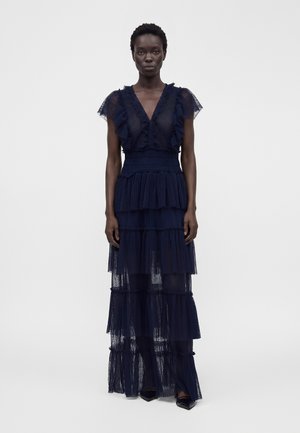 Femme debout portant une robe longue bleu marine en dentelle à volants avec des manches à volants et un col en V, sur un fond clair uni.