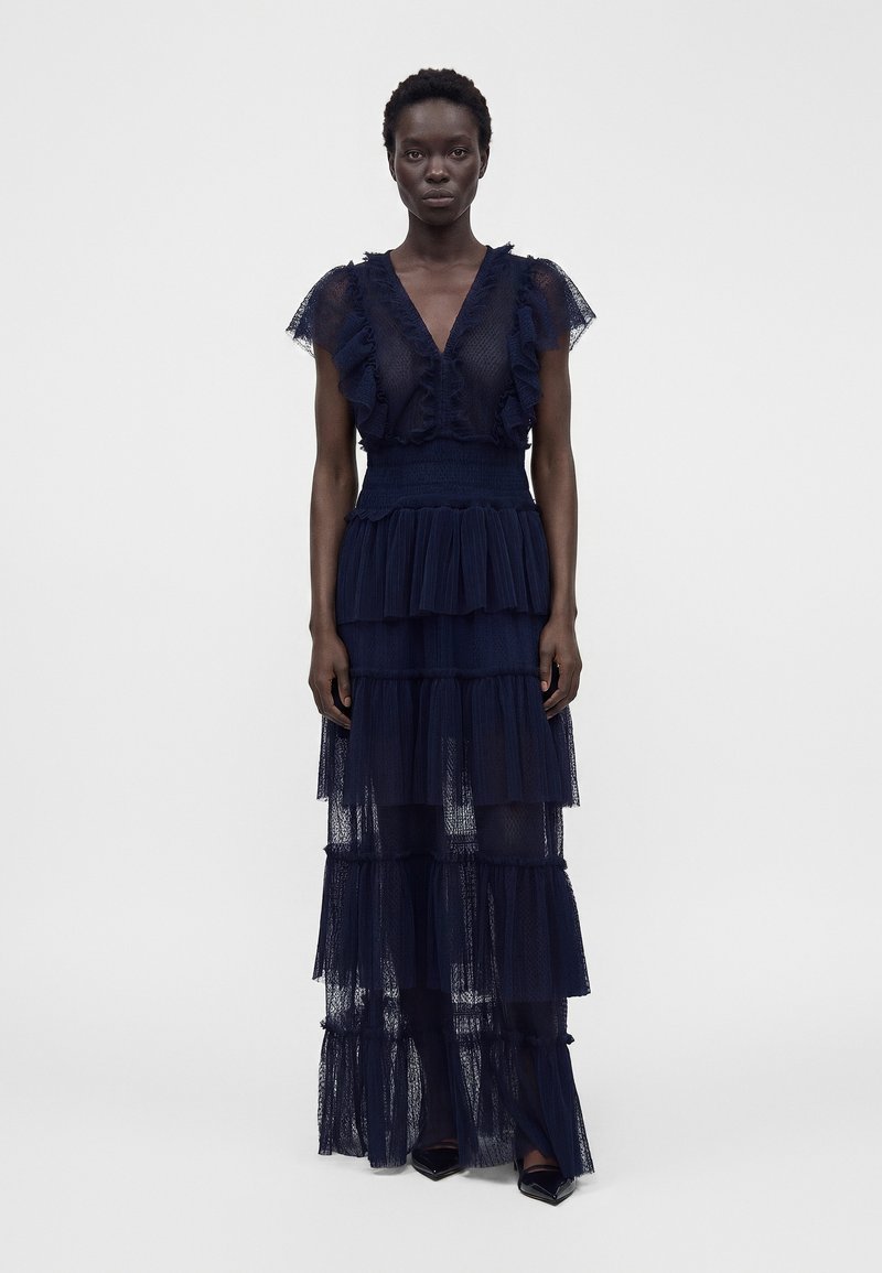 Femme debout portant une robe longue bleu marine en dentelle à volants avec des manches à volants et un col en V, sur un fond clair uni.