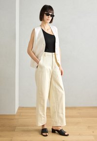 Lichtbeige mouwloze vest met knopen, zwarte tanktop, crèmekleurige wijde broek en zwarte open teen muiltjes op een houten vloer tegen een grijze muur.