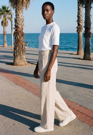 Tânără în cămașă albă, pantaloni bej cu croială largă și adidași albi mergând pe o promenade la malul mării, marginită de palmieri.