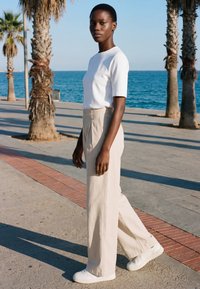 Jeune femme en chemise blanche, pantalon beige à jambes larges et baskets blanches marchant sur une promenade en bord de mer bordée de palmiers.