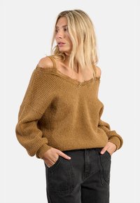 Maglione oversized a spalle scoperte in tessuto di maglia marrone testurizzato con polsini a coste, indossato con jeans di denim neri a vita alta.