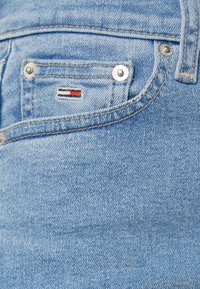 Vaqueros de denim azul claro con un pequeño bolsillo delantero que presenta un parche de logo tricolor y botones de presión plateados. Textura suave y diseño clásico.
